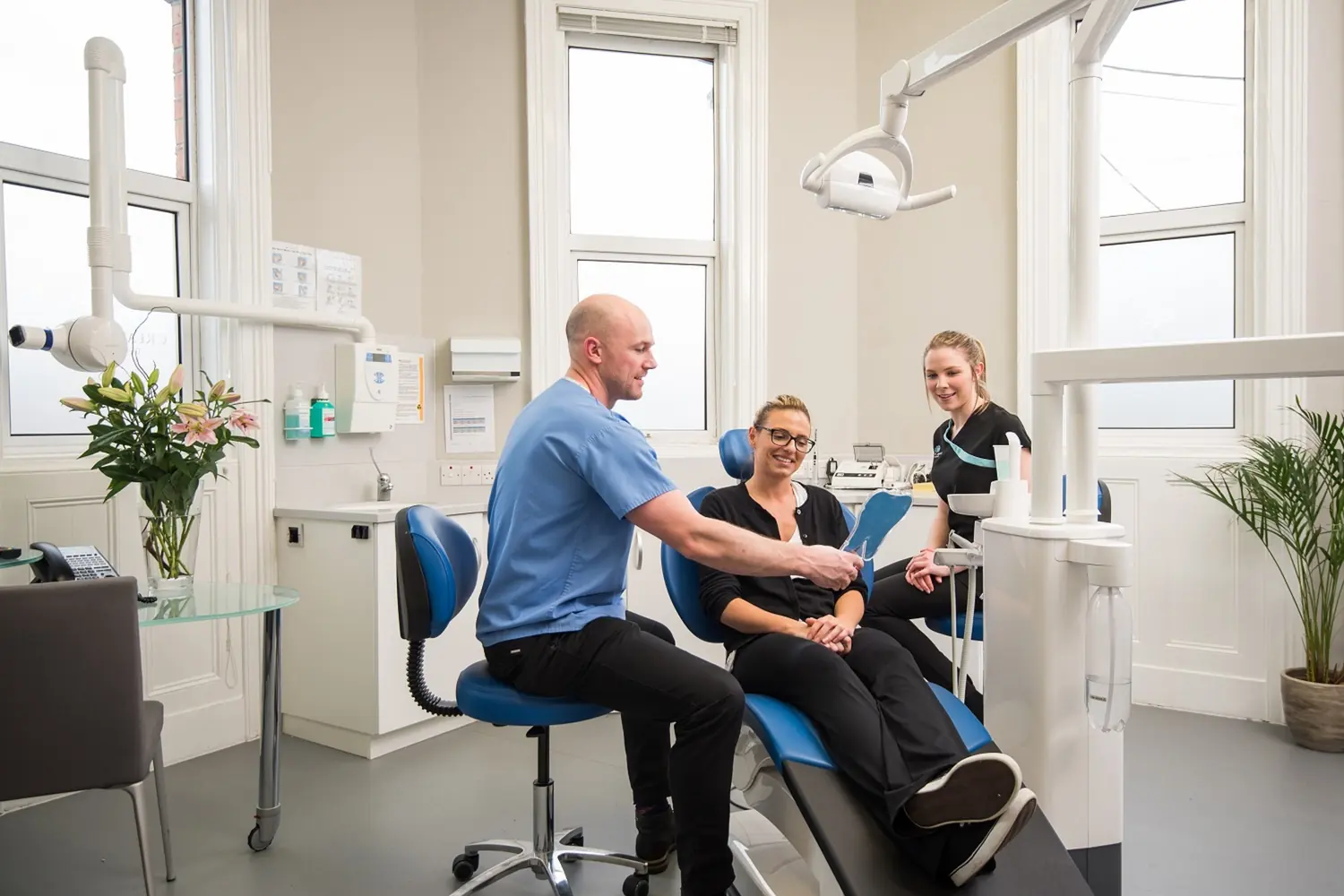 A dentist explaining a treatment plan to a smiling patient in a bright exam room with blue chairs